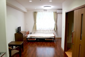窓採光の明るい個室。ベッド、テレビ、机が配置された居住空間